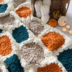 May include: A handmade crochet blanket in a patchwork design. The blanket features squares in shades of cream, teal, orange, and taupe. The blanket is next to a basket with yarn, mini pumpkins, and candles.