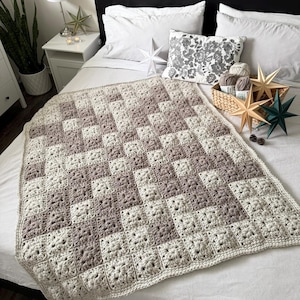Pode incluir: Um cobertor de crochê feito à mão em tons de creme e taupe, com um padrão quadrado. Exibido em uma cama adornada com estrelas decorativas e uma almofada floral.
