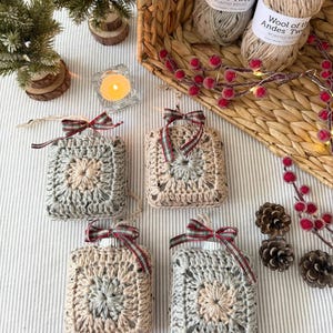 Pode incluir: Quatro enfeites quadrados de crochê em tons de cinza e bege, cada um com uma fita xadrez. Uma cesta contém fios com a etiqueta "Wool of the Andes Tweed". A cena inclui pequenas árvores de Natal, uma vela e pinhas.