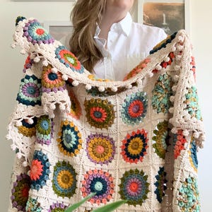 Pode incluir: Um cobertor de crochê creme com padrões quadrados florais coloridos. O cobertor apresenta quadrados em tons de laranja, azul, verde e roxo. As bordas são finalizadas com uma borda ondulada. O cobertor é segurado, mostrando seu design.