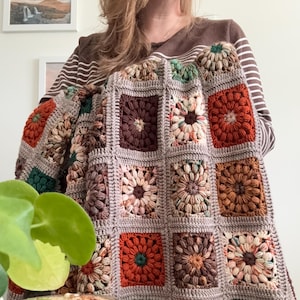 May include: A handmade crochet blanket featuring a patchwork design with square floral motifs in shades of brown, orange, green, and cream. The blanket is held up, showcasing its intricate texture and color palette, perfect for home decor.