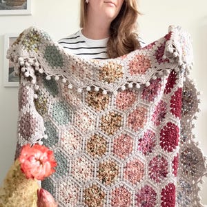 May include: A handmade crochet blanket featuring a hexagonal pattern in shades of pink, beige, and green. The blanket has a scalloped edge with small pom-poms. The blanket is displayed in front of a person wearing a striped shirt.