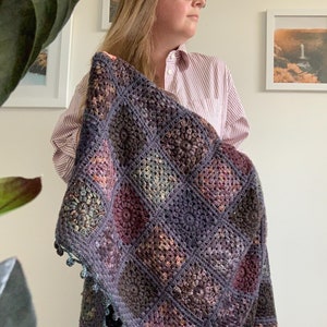 May include: A crocheted blanket with a square pattern in shades of purple, blue, and green. The blanket has a fringe border with pom-poms.