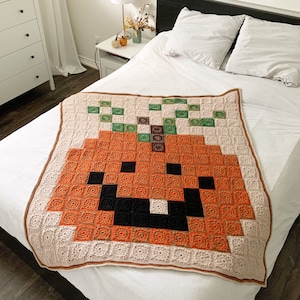 Peut inclure: Une couverture au crochet avec un motif de citrouille pixelisé dans des tons d'orange, de brun et de noir. La couverture est posée sur un lit blanc avec des oreillers.