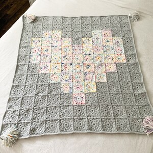 Könnte beinhalten: Eine graue Häkeldecke mit einem bunten Herzdesign in der Mitte. Die Decke hat Fransen an den Ecken.