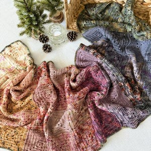 Op de afbeelding: Een display van drie kleurrijke, gehaakte dekens in tinten roze, paars en groen. De dekens zijn opgevouwen en gedrapeerd, met een gevlochten mand en kleine decoratieve boompjes op de achtergrond. De dekens zijn handgemaakt.