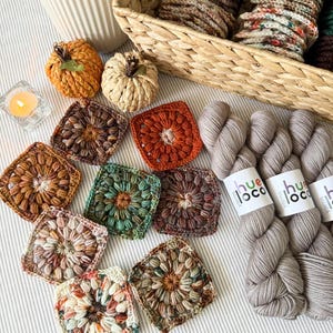 May include: An overhead shot features a collection of handmade crochet items. There are several colorful granny squares, two small crochet pumpkins, and skeins of gray yarn. A basket filled with yarn and a lit candle are also present, creating a cozy, autumnal theme.