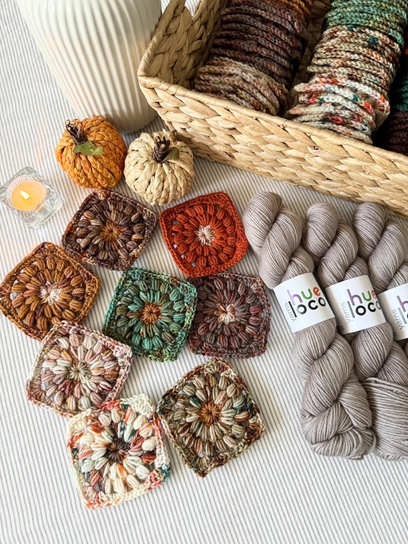 May include: An overhead shot features a collection of handmade crochet items. There are several colorful granny squares, two small crochet pumpkins, and skeins of gray yarn. A basket filled with yarn and a lit candle are also present, creating a cozy, autumnal theme.