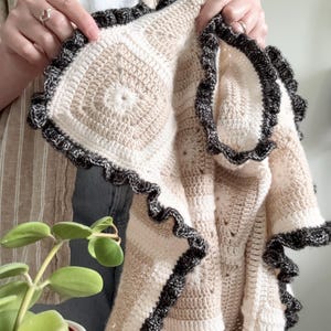 Puede incluir: Una manta de ganchillo hecha a mano con una serie de motivos cuadrados en tonos crema y beige. La manta está rematada con un borde de volantes en gris carbón oscuro que crea un borde texturizado alrededor de todo el perímetro de la pieza.
