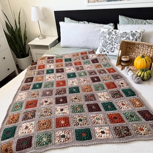 May include: A handmade crochet blanket with a floral pattern in shades of brown, green, orange, and cream. The blanket is displayed on a bed with white bedding. The blanket's border is a solid gray color.