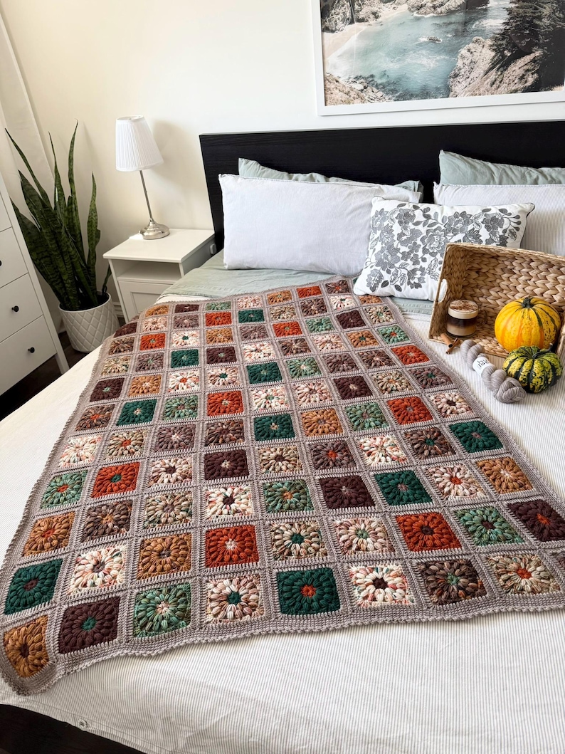May include: A handmade crochet blanket with a floral pattern in shades of brown, green, orange, and cream. The blanket is displayed on a bed with white bedding. The blanket's border is a solid gray color.