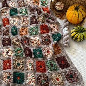 May include: A handmade, square crochet blanket in a variety of fall colors, including rust, green, and brown. The blanket is draped over a surface with a woven basket, a small pumpkin, and a ball of yarn.