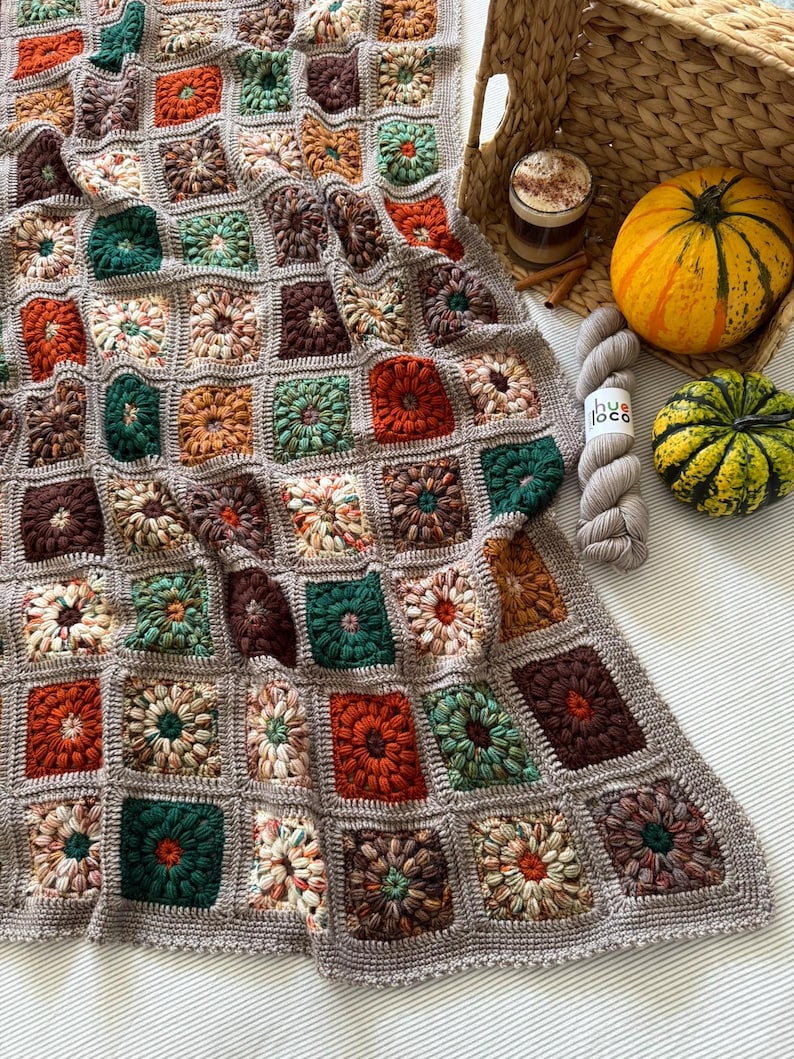 May include: A handmade, square crochet blanket in a variety of fall colors, including rust, green, and brown. The blanket is draped over a surface with a woven basket, a small pumpkin, and a ball of yarn.