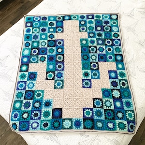 Può includere: Una coperta all'uncinetto con un motivo geometrico di quadrati blu e verdi. La coperta è realizzata con filato beige e ha un bordo blu scuro.