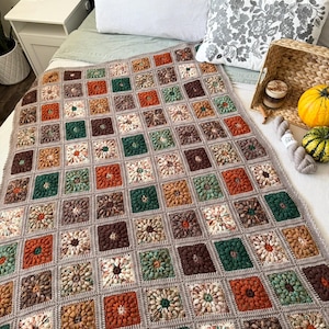 May include: A handmade crochet blanket in a patchwork design. The blanket features squares in shades of brown, orange, green, and cream, with a gray border. The blanket is displayed on a bed with pillows and decorative items.