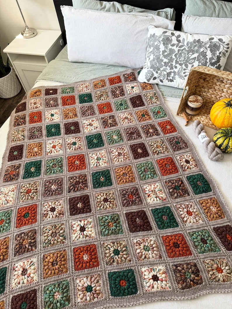 May include: A handmade crochet blanket in a patchwork design. The blanket features squares in shades of brown, orange, green, and cream, with a gray border. The blanket is displayed on a bed with pillows and decorative items.