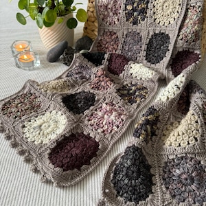 May include: A handmade crochet scarf in a granny square pattern. The scarf features squares in shades of gray, cream, and burgundy, with a border of small bobbles. The scarf is displayed on a striped surface.