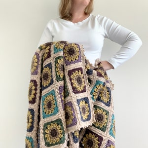May include: A crocheted throw blanket with a square pattern featuring colourful flowers. The blanket is made with a variety of yarns in shades of brown, beige, green, blue, and purple.