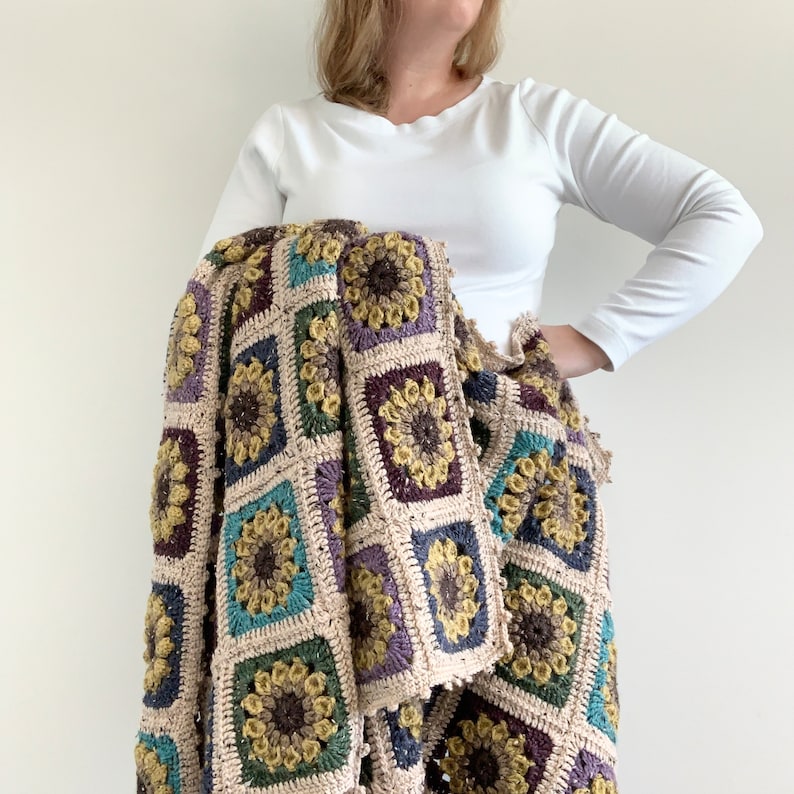 May include: A crocheted throw blanket with a square pattern featuring colourful flowers. The blanket is made with a variety of yarns in shades of brown, beige, green, blue, and purple.