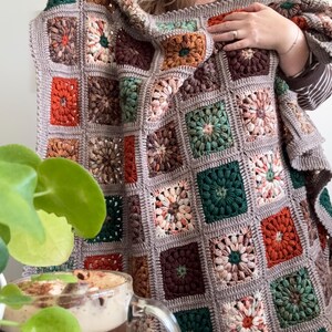 May include: A handmade, square-patterned crochet blanket in a variety of colors, including orange, green, brown, and cream. The blanket is held up, showcasing its intricate design and cozy texture. The blanket is approximately 60 inches by 60 inches.