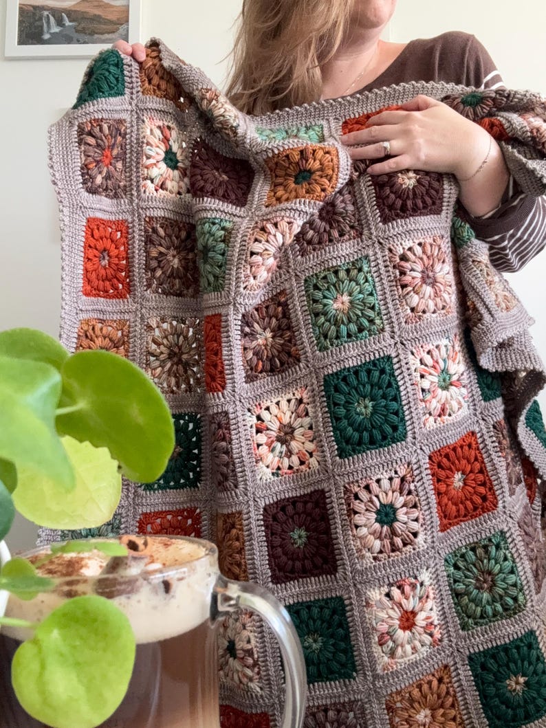 May include: A handmade, square-patterned crochet blanket in a variety of colors, including orange, green, brown, and cream. The blanket is held up, showcasing its intricate design and cozy texture. The blanket is approximately 60 inches by 60 inches.
