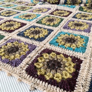 May include: A crocheted throw blanket with a square pattern featuring a sunflower design in shades of brown, yellow, purple, blue, and green.