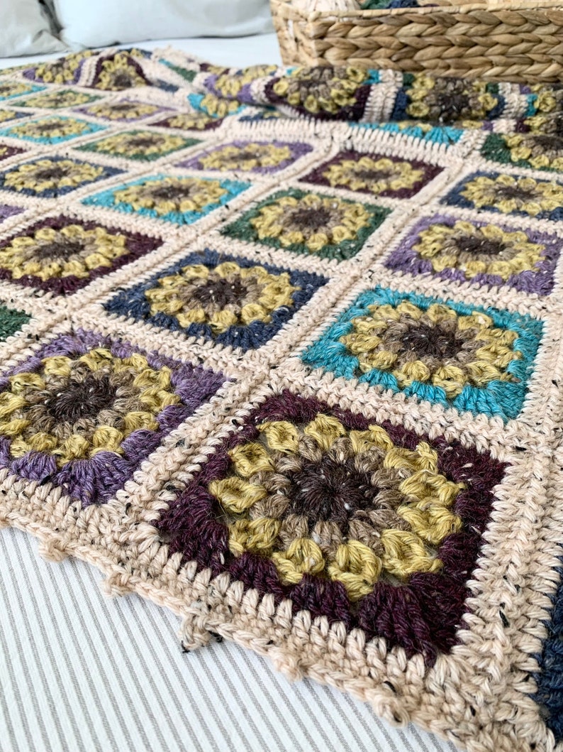 May include: A crocheted throw blanket with a square pattern featuring a sunflower design in shades of brown, yellow, purple, blue, and green.