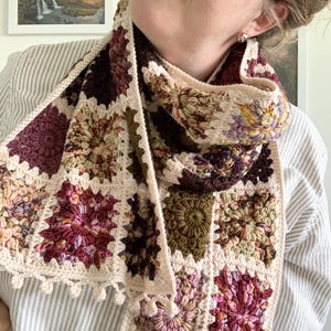 May include: A crocheted scarf with a cream background and colorful floral patterns in shades of pink, purple, green, and brown. The scarf has a fringe border and is made with a granny square stitch.