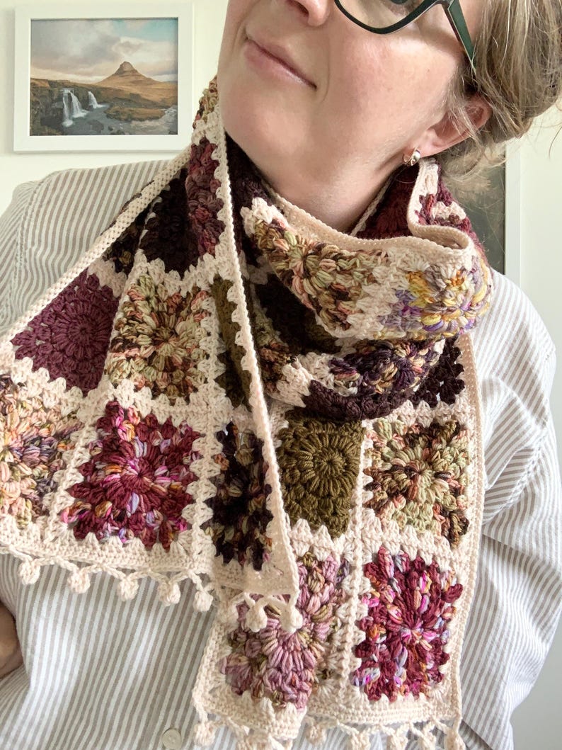 May include: A crocheted scarf with a cream background and colorful floral patterns in shades of pink, purple, green, and brown. The scarf has a fringe border and is made with a granny square stitch.