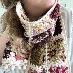 May include: A crocheted scarf with a variety of colorful squares. The scarf is made with a cream-colored yarn and features different colored flowers in each square. The scarf has a fringe on one end.