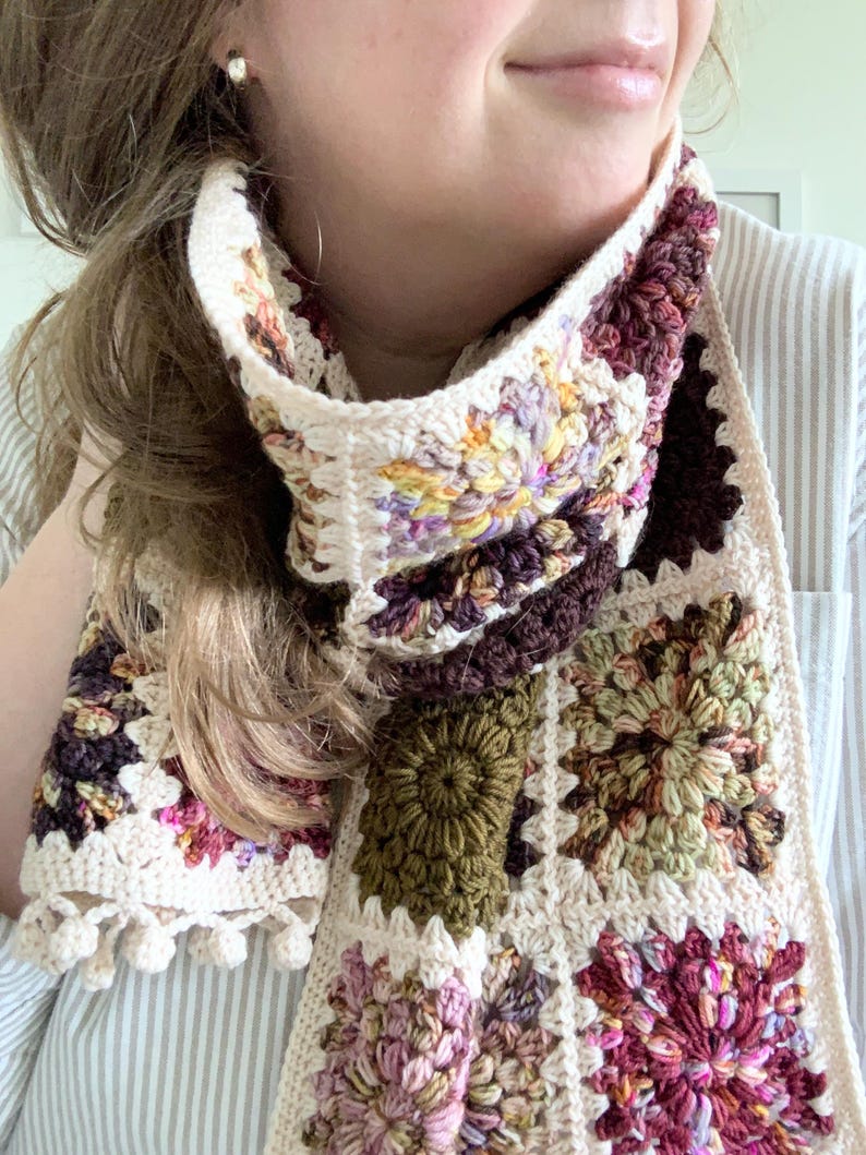 May include: A crocheted scarf with a variety of colorful squares. The scarf is made with a cream-colored yarn and features different colored flowers in each square. The scarf has a fringe on one end.