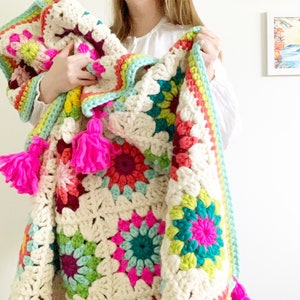 May include: A handmade, colorful crochet blanket featuring a granny square design. The blanket showcases a cream-colored base with vibrant squares in shades of pink, teal, red, and yellow. Bright pink tassels adorn the corners, adding a playful touch to this cozy, decorative item.