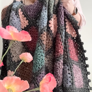以下が含まれることがあります： 手作りの、マルチカラーのかぎ針編みの毛布。花柄です。グレー、紫、ピンク、茶色の正方形が特徴です。縁は濃いグレーで、小さなポンポンが付いています。ピンクの花が前景にあります。