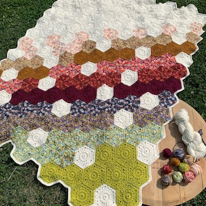 May include: A crocheted blanket with a hexagonal pattern in shades of green, white, purple, red, brown, and pink. The blanket is laid out on green grass with a wooden tray holding balls of yarn in various colours.