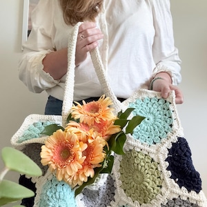 May include: A handmade crochet tote bag constructed from joined hexagonal motifs in shades of navy blue, sage green, light blue, and grey, outlined in cream yarn. The bag features two sturdy cream-colored crochet handles and holds orange flowers.