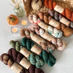 May include: A collection of colorful yarn skeins in shades of brown, orange, and green, arranged with decorative pumpkins, candles, and a woven basket. The yarn is labeled "Pumpkin Spice Everything". The scene evokes a cozy, autumnal theme.