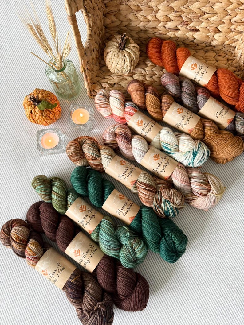 May include: A collection of colorful yarn skeins in shades of brown, orange, and green, arranged with decorative pumpkins, candles, and a woven basket. The yarn is labeled "Pumpkin Spice Everything". The scene evokes a cozy, autumnal theme.