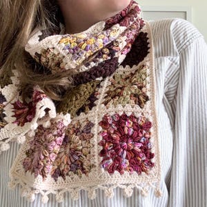 May include: A crocheted scarf with a multi-colored granny square pattern. The scarf is made with a variety of shades of brown, pink, green, and cream yarn. The scarf has a crocheted fringe along the bottom edge.