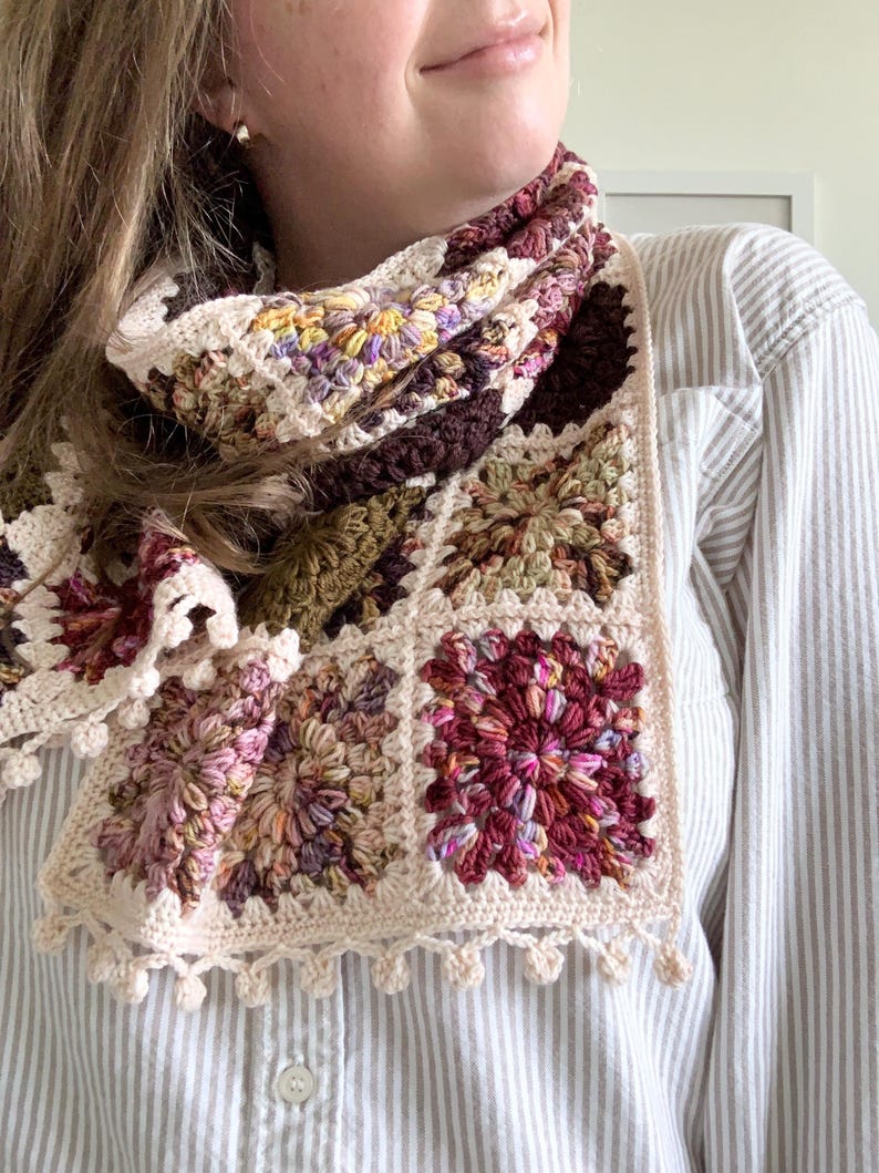 May include: A crocheted scarf with a multi-colored granny square pattern. The scarf is made with a variety of shades of brown, pink, green, and cream yarn. The scarf has a crocheted fringe along the bottom edge.