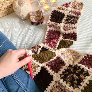 May include: A person is crocheting a blanket with a variety of colors, including brown, green, and cream. The blanket is made of squares with a floral pattern. The person is using a red crochet hook and a ball of yarn.