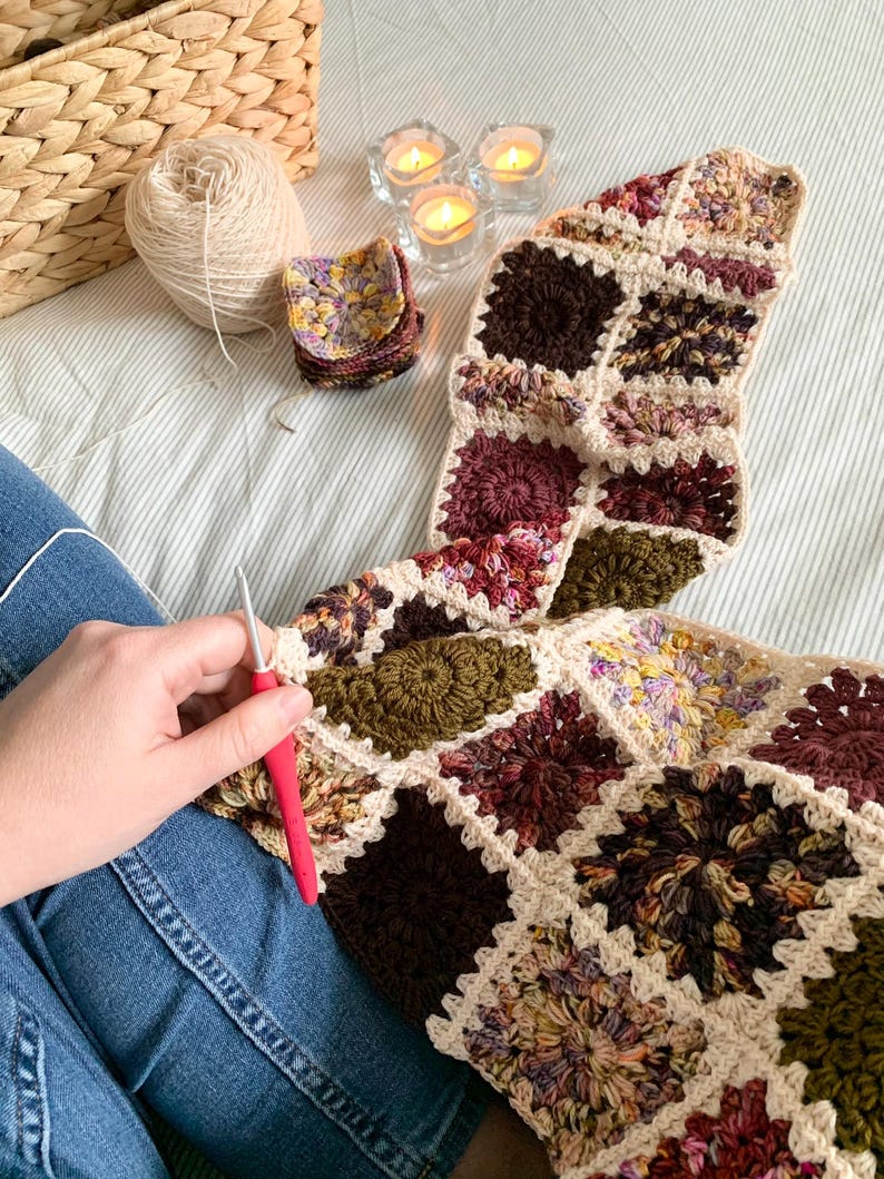 May include: A person is crocheting a blanket with a variety of colors, including brown, green, and cream. The blanket is made of squares with a floral pattern. The person is using a red crochet hook and a ball of yarn.