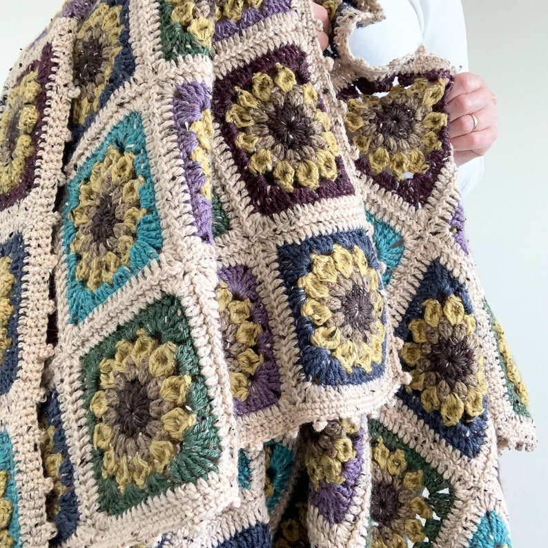 May include: A crocheted throw blanket with a patchwork design featuring squares of different colours and textures. The blanket is made with a variety of yarns in shades of brown, green, purple, and blue. The blanket is folded and draped over a person's arm.