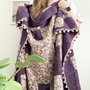 以下が含まれることがあります： 紫とクリーム色の毛糸で編まれたブランケット。縁は編み込みになっており、ポンポンが付いています。ブランケットは折り畳まれ、人の腕にかけられています。