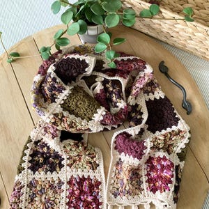 May include: A crocheted scarf with a granny square pattern in shades of brown, green, and pink. The scarf has a white crocheted border with a fringe.