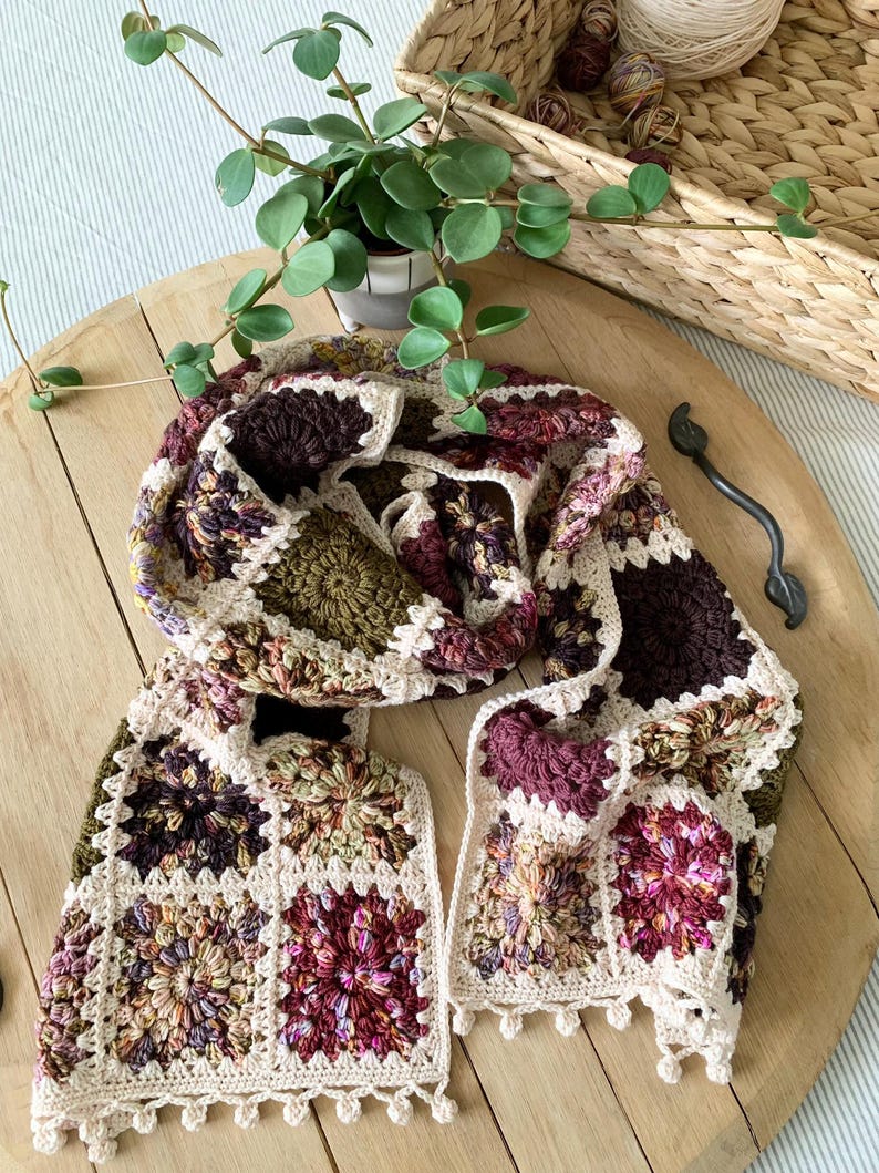 May include: A crocheted scarf with a granny square pattern in shades of brown, green, and pink. The scarf has a white crocheted border with a fringe.
