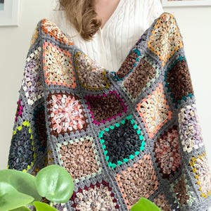 May include: A handmade, colorful crochet blanket. The blanket features a patchwork design with square motifs in various colors, including gray, brown, pink, and teal. The blanket is made of soft yarn and is perfect for snuggling up on the couch.