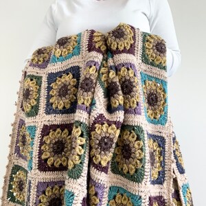 May include: A crocheted throw blanket featuring a sunflower design in shades of brown, green, purple, and blue. The blanket is crafted with a mix of textures and has a fringed border.