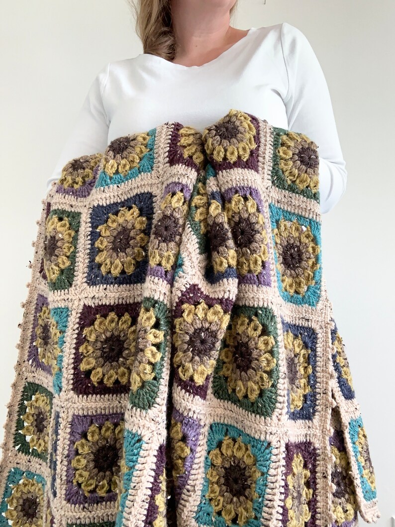 May include: A crocheted throw blanket featuring a sunflower design in shades of brown, green, purple, and blue. The blanket is crafted with a mix of textures and has a fringed border.