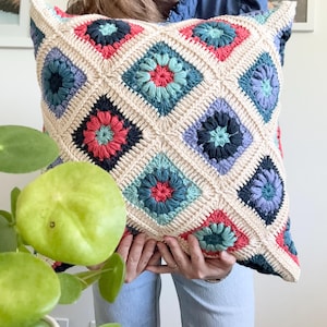 May include: A square crochet pillow cover featuring a repeating granny square pattern in cream, coral pink, teal, and various shades of blue. The design consists of interconnected diamond-shaped motifs with a central floral-like stitch detail.