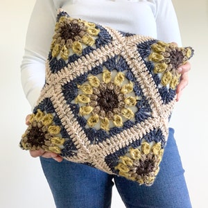 May include: A crocheted pillow with a square pattern featuring brown, beige, and yellow flowers. The pillow is held by a person wearing blue jeans.